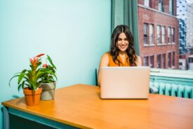 Confident woman in a bright office working on a laptop, surrounded by urban scenery and greenery.